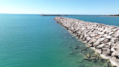 Rockwall-jetty-at-harbor-entrance-with-crystal-blue-water,-aerial-dolly