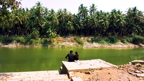 beautiful-couple-setting-on-beautiful-Lake-Step-of-palm-forest-area,-aerial-camera-moving-zoom-out-to-a-beautiful-couple