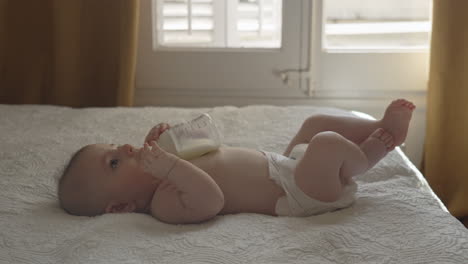 Newborn-self-feeding-milk-bottle-playful-mood-in-white-sunny-bedroom,-cozy-baby