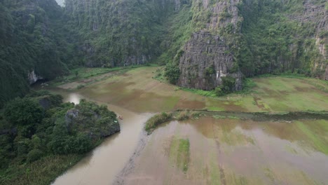 Flooded-Rice-Fields-On-The-Riverbanks-of-Ngo-Dong-River-in-Ninh-Binh-Vietnam,-People-Cruising-on-Boats-In-Limestone-Mountain-Gorge---Drone-View
