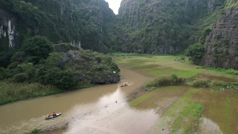 Sampan-Tour-Boats-With-Tourists-Cruise-on-Ngo-Dong-River-Running-in-Limestone-Ravine-in-Ninh-Binh-National-Park,-Flooded-Rice-Fields-On-Riverbanks---aerial-ascending