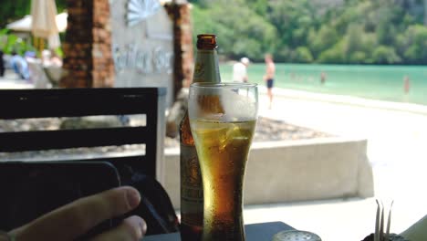 Kaltes-Bier-Auf-Einem-Tisch-Mit-Einem-Weißen-Sandstrand-Im-Hintergrund