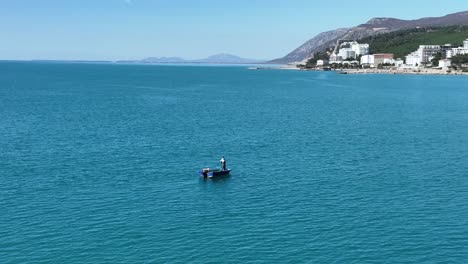 Solo-fisherman-pulls-net-from-boat-in-open-ocean,-coastal-town-in-view