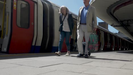Elderly-Couple-Walking-Along-Platform-Pulling-Wheeled-Luggage-At-Harrow-Past-Met-Line-Train-As-It-Departs