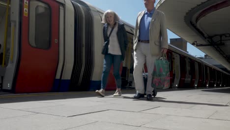 Pareja-De-Ancianos-Caminando-A-Lo-Largo-De-La-Plataforma-Tirando-De-Equipaje-Con-Ruedas-En-Grada-Pasando-El-Tren-De-La-Línea-Met-A-Medida-Que-Sale