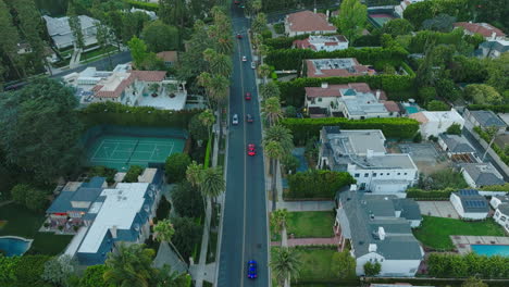 Volando-Con-El-Auto,-Dron-Sobre-La-Carretera-De-Beverly-Hills-Mientras-Un-Elegante-Auto-Rojo-Conduce-Por-Propiedades-De-Lujo