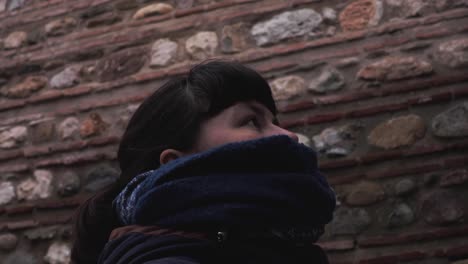 Close-shot-of-a-brunette-young-girl-in-winter-clothes-standing-next-to-a-maroon-brick-wall-of-an-old-building,-looking-upwards