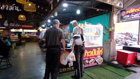 2-Hombres-Turistas-Comprando-Comida-En-Un-Mercado-Interior-De-Alimentos-Mientras-Sostienen-Cascos-De-Moto