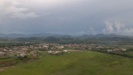 Descending-Drone-Footage-of-Small-Town-with-Lush-Greenery-and-Distant-Mountains-under-Heavy-Clouds