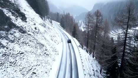 Aerial-close-view-of-a-small-car-driving-on-a-snow-covered-mountain-road-surrounded-by-forests-during-a-cloudy-winter-day-with-snowfall-in-Switzerland