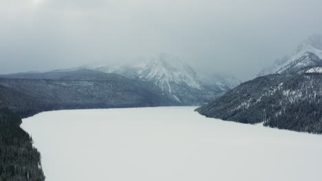 Aerial-moving-over-frozen-lake-with-large-mountains-covered-in-clouds-behind
