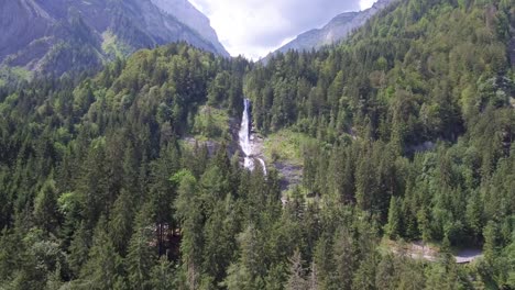 Drone-flight-over-a-lush-green-alpine-forest-approaching-a-glacier-melt-cascading-down-the-rock-face