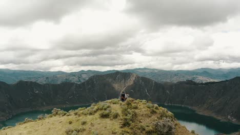 Ascending-tracking-shot-of-couple-standing-on-peak-of-mountain-and-enjoying-volcano-crater-lake-view-during-cloudy-day