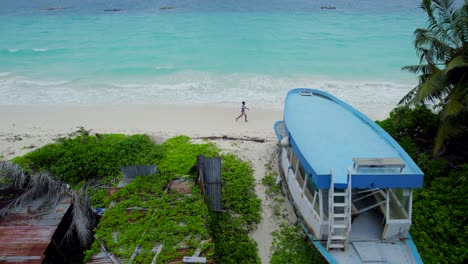 Aerial-drone-view-of-Maldivian-man-walking-through-tropical-beach-next-to-a-wrecked-ship
