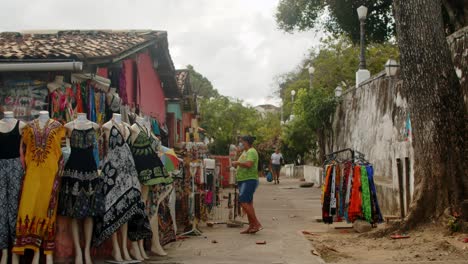 Pequeña-Tienda-De-Ropa-Callejera-Llena-De-Vestidos-Coloridos-Con-Una-Anciana-Con-Máscara-En-Un-Día-Nublado