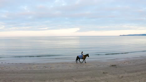 A-young-beautiful-brunette-girl-with-long-hair-in-a-blue-flowing-dress-rides-a-brown-horse-on-Donabate-beach,-Ireland
