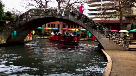 Die-Flussboote-Auf-Dem-San-Antonio-Riverwalk-Fahren-Ständig-Durch-Die-Innenstadt,-Deren-Bunte-Lichter-Auf-Der-Oberfläche-Zu-Tanzen-Scheinen