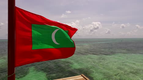 Maldivian-flag-waving-over-a-platform-floating-on-shallow-lagoon-near-tropical-island-with-a-cloudy-sky-background,-copy-space