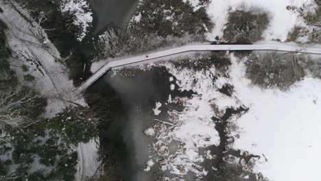 Aerial-view-of-person-running-over-wooden-path-next-to-frozen-lake-with-forest-covered-in-snow