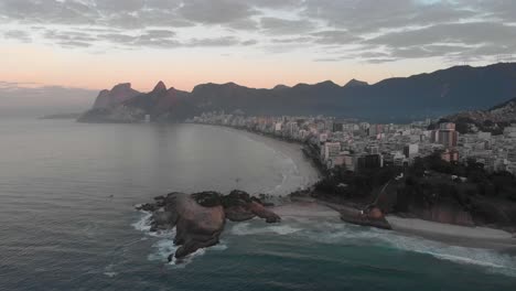 Aerial-sunrise-of-Rio-de-Janeiro-with-Arpoador-in-the-foreground-and-Ipanema-beach-and-wider-cityscape-in-the-background-with-clouds-over-the-city-at-early-morning