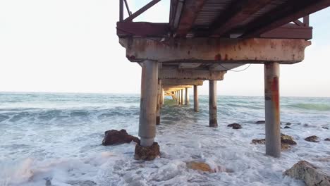 drone-flying-under-wooden-pier-in-marbella-malaga-spain,-beautiful-provoking-shot