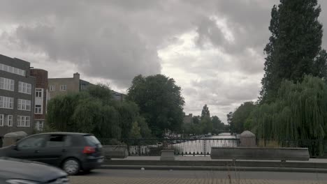 Eine-Traurige-Aufnahme-Der-Amsterdamer-Kanäle,-Vorbeifahrender-Menschen-Und-Autos,-Große-Graue-Wolken-Im-Hintergrund