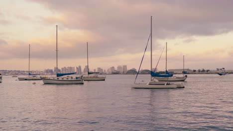 Sailboats-on-water-in-San-Diego-California-harbor-during-pink-cloudy-sunset,-Coronado-island-with-navy-buildings-and-downtown-in-the-background