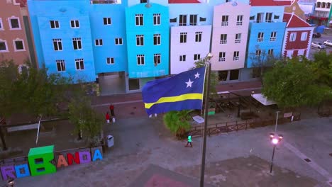 Curacao-flag-on-pole,-colorful-buildings-of-Otrobanda,-Willemstad-in-background,-aerial-shot-at-dusk