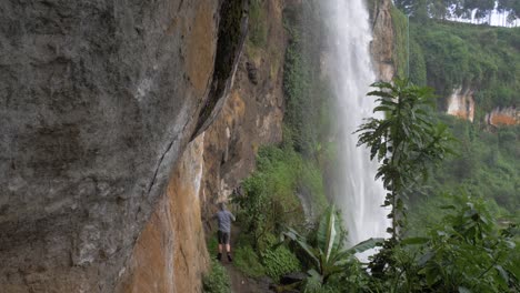 Slow-motion-shot-of-a-hiker-walking-along-a-small-cliff-trail-underneath-a-tropical-waterfall
