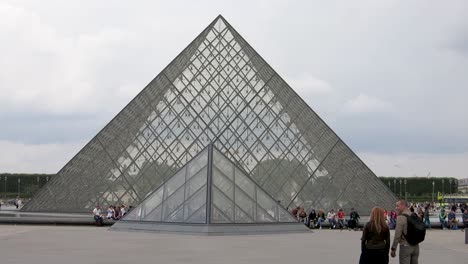Pirámide-Del-Louvre-Con-Turistas-Sentados-En-Su-Base,-París,-Francia