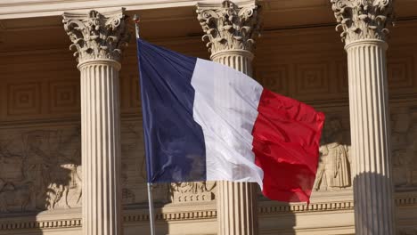 Bandera-Francesa-Moviéndose-En-El-Viento-Frente-A-Un-Edificio-Inspirado-En-Las-Antiguas-Columnas-De-Grecia-Con-Su-Sombra-En-Una-Pared
