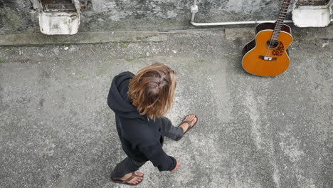Slow-motion,-top-shot-of-a-young-man-wearing-a-black-hoodie-walking-towards-a-classical-guitar,-then-taking-it-with-a-hand-and-holding-it-still,-in-an-abandoned-structure,-on-a-cloudy-day