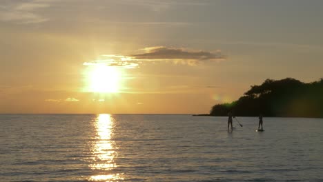 Majestic-large-bright-sun-in-the-horizon-illuminating-the-ocean-during-sunset-with-two-people-paddle-boarding-in-the-distance