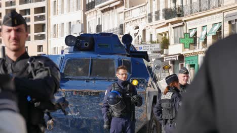 Ein-Polizist-In-Voller-Kampfmontur-Beobachtet-Eine-Menge-Demonstranten-Mit-Gelben-Jacken,-Während-Ein-Blauer-Polizeipanzer-Seine-Wasserwerfer-Ein-Paar-Grad-In-Richtung-Kamera-Bewegt