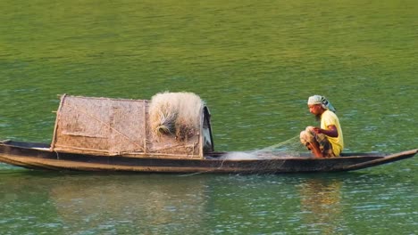 A-skilled-fisherman-navigates-a-traditional-wooden-boat-on-the-tranquil-waters-of-Surma-river,-deftly-casting-his-fishing-net-to-catch-a-variety-of-fish