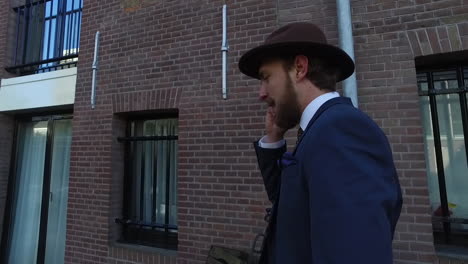 Ein-Eleganter-Mann-Im-Dandy-Stil-Mit-Blauem-Anzug-Und-Bart-Telefoniert,-Während-Er-In-Amsterdam-Gegen-Eine-Backsteinmauer-Läuft
