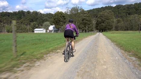 Woman-with-pony-tail-on-a-mountain-bike-coasting-on-a-gravel-road-with-a-farmhouse-in-the-distance