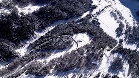 Aerial-view-of-the-Bernina-Express-panorama-train-going-through-a-snow-covered-mountain-winter-landscape-with-forests-on-a-sunny-day-in-Alp-Grum,-Switzerland