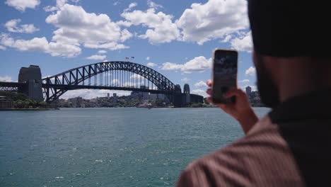 Punjabi-Sikh-Hombre-Con-Smartphone-Tomando-Fotos-Del-Puente-Del-Puerto-De-Sydney-En-Nsw,-Australia