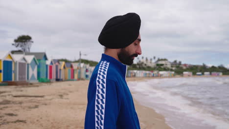 Seitenansicht-Eines-Indischen-Mannes-Mit-Turban,-Der-Schaut-Und-Denkt,-Während-Er-Am-Strand-Von-Dendy-Street-Mit-Badeboxen-In-Brighton-In-Australien-Steht