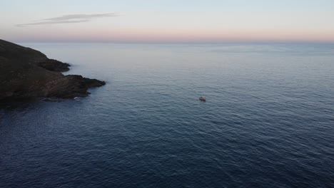 Aerial-drone-view-of-Cornish-coast-at-dusk,-featuring-a-small-fishing-boat-and-enchanting-scenery