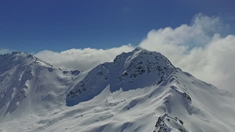 White-snow-covered-mountain-slopes-with-fluffy-clouds-in-sky,-aerial-view