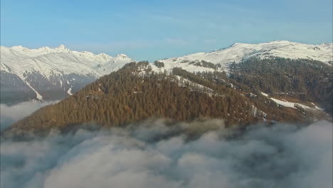 Mountain-covered-in-forest-and-snow,-flying-above-clouds-view