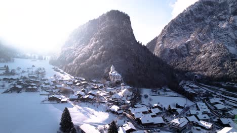 Aerial-view-of-a-mountain-village-with-a-church-and-snow-covered-mountains-in-Switzerland-on-a-sunny-winter-day