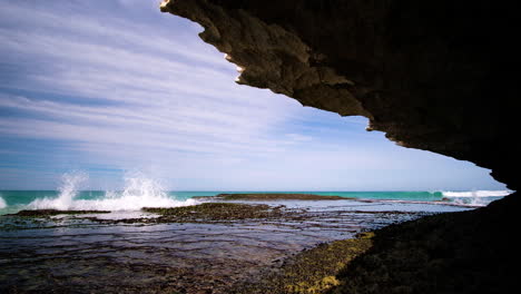 Small-wave-breaking-on-rocks-on-sunny-day,-shot-from-a-cave,-static-shot