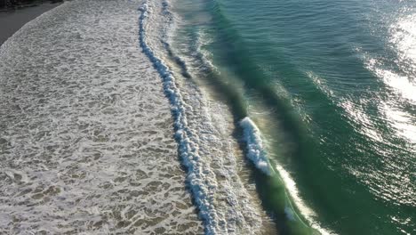 Wunderschöne-Luftaufnahme-Eines-Urlaubsziels-Mit-Blauem-Meer,-Sanften-Wellen-Und-Surfern-Im-Hintergrund-In-Der-Nähe-Eines-Weißen-Sandstrandes