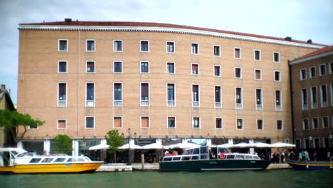Breiter-Blick-Auf-Die-Straße-In-Venedig,-Italien,-Wie-Boote-Auf-Der-Straße-Vor-Einem-Gebäude-Mit-Einem-Restaurant-Andocken,-Während-Menschen-Und-Touristen-Vorbeigehen