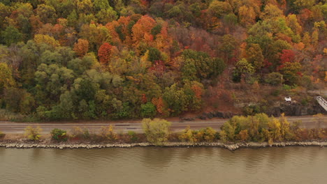 aerial-drone-shot-over-the-Hudson-River,-while-the-camera-trucks-right,-focusing-on-the-autumn-colored-tree-tops,-roadway---Metro-North's-train-tracks-on-a-cloudy-evening-in-Beacon,-NY