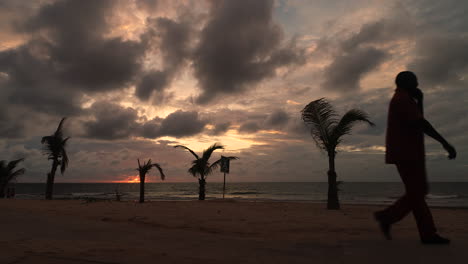 Afrikanischer-Sillouette-Mann-Mit-Handy-Geht-Bei-Sonnenuntergang-Am-Strand-Von-Senegambia,-Serekunda,-Gambia-Vorbei