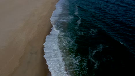 smooth-aerial-flight-over-the-waters-edge-on-a-sandy-beach-with-breaking-waves